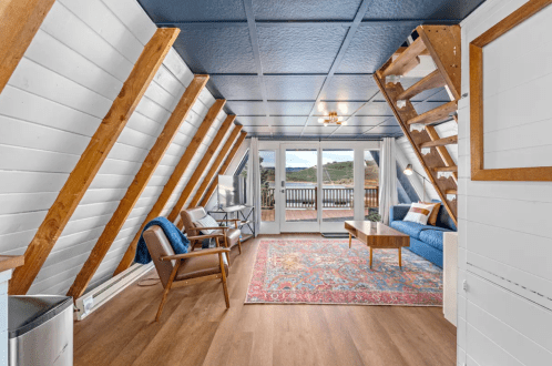 Cozy attic living space with wooden beams, a blue sofa, coffee table, and large windows overlooking a scenic view.